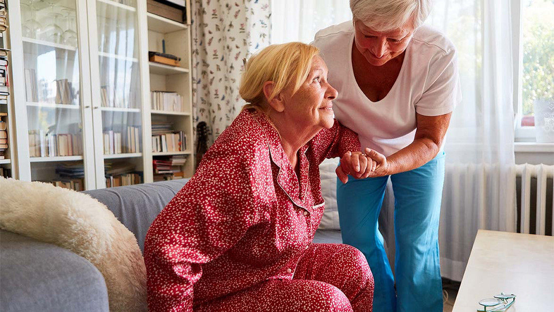 Eine Pflegekraft hilft einer älteren Frau beim Aufstehen vom Sofa. 