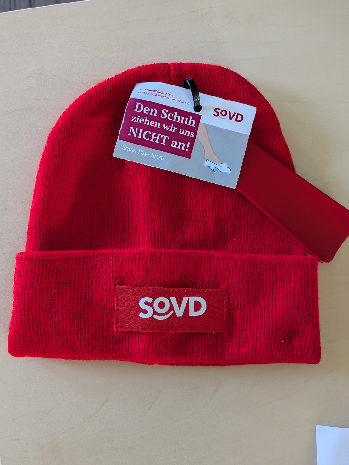 Rote Mütze mit SoVD-Logo und Visitenkarte "iesen Schuh ziehen wir uns nicht an! - Equal Pay Jetzt!"