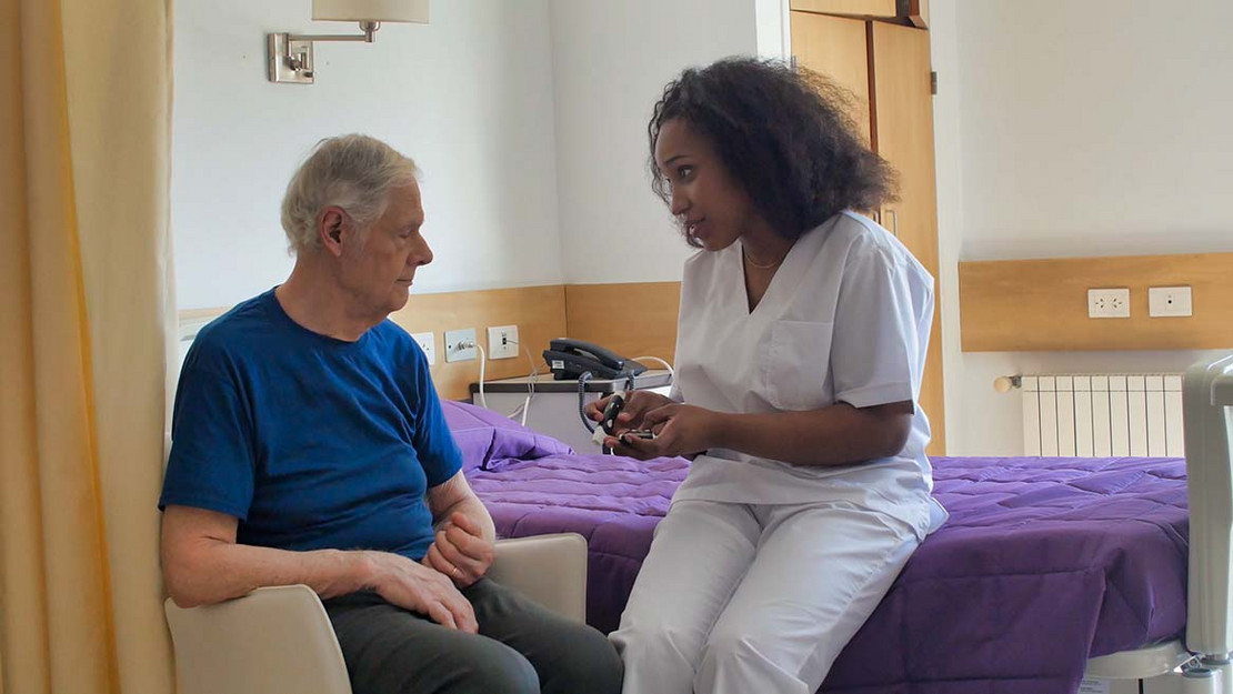 Pflegerin sitzt auf dem Bett und spricht mit einem Heimbewohner.